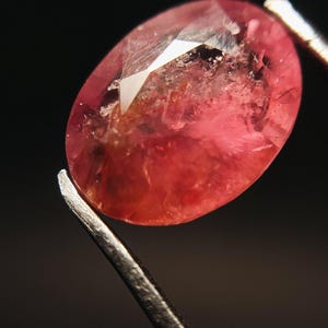 Puede incluir: Una gema facetada de forma ovalada, sujeta con pinzas. La gema es de color rosa vibrante con inclusiones internas y una superficie reflectante. El fondo oscuro resalta el color y la claridad de la gema.