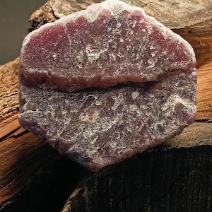 Könnte beinhalten: Ein grob geschliffener, roher Rubin-Edelstein mit tiefroter bis violetter Farbe. Der Stein hat eine strukturierte Oberfläche und wird auf verwittertem Holz präsentiert, was seine natürliche Schönheit hervorhebt. Ein Unikat für Schmuckherstellung oder Sammler.