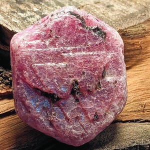 Könnte beinhalten: Ein roher, ungeschliffener Rubin-Edelstein mit tiefrosa Farbton und dunklen Einschlüssen. Der Naturstein hat eine strukturierte Oberfläche und wird auf einem Holzgrund präsentiert, der seine rohe, organische Form zeigt. Ein schönes Beispiel für natürliche Mineralien.
