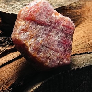 Può includere: Un grezzo rubino non tagliato, di colore rosso intenso e rosa. La superficie della pietra ha un aspetto strutturato, con visibili strutture cristalline. Il rubino è posto su uno sfondo di legno scuro e strutturato.