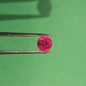 Puede incluir: Una gema redonda de color rojo vibrante sostenida por unas pinzas plateadas sobre un fondo verde. La gema es de un color profundo y saturado, y las pinzas están bien enfocadas. La imagen está bien iluminada, resaltando el brillo de la gema.