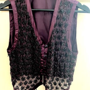May include: A plum-coloured vest with a V-neck and three buttons. The vest features black beaded embellishments in a geometric pattern on the front panels and a lace trim at the bottom. The vest is hanging on a black hanger.