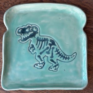 May include: A turquoise ceramic plate shaped like a slice of bread, with a dark blue dinosaur skeleton design. The plate has rounded edges and a smooth surface. Ideal for serving snacks or small meals, it measures approximately 20cm x 20cm.