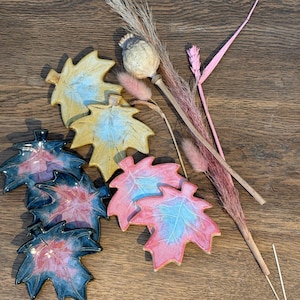 May include: Set of six ceramic maple leaf dishes in three colours: yellow, pink, and black. Each leaf has a blue glaze detail. The dishes are arranged on a wooden surface with dried flowers.