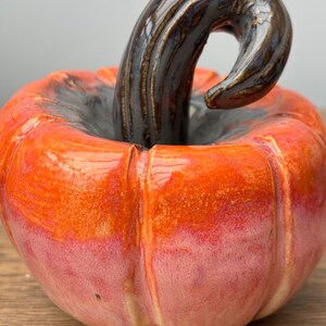 May include: A ceramic pumpkin with a glossy orange and pink glaze. The pumpkin has a detailed stem with a dark brown glaze.