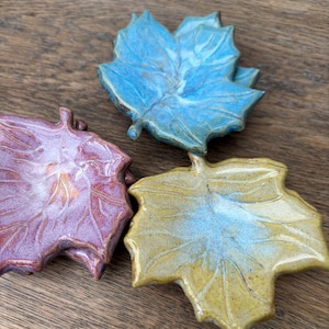 May include: Three ceramic leaf-shaped dishes in different colours. The dishes are stacked on top of each other. The top dish is blue, the middle dish is purple, and the bottom dish is yellow.