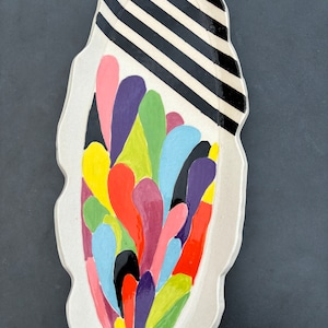 May include: A white ceramic serving dish with a black and white striped design on one side and a colourful abstract design on the other side. The abstract design features a variety of colours, including red, orange, yellow, green, blue, purple, and pink.