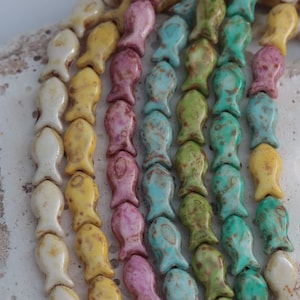 May include: Close-up of colourful ceramic fish beads in white, yellow, pink, blue, and green. The beads are strung together, creating a textured pattern. The beads are arranged on a white, textured surface.