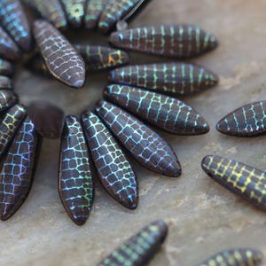 以下が含まれることがあります： クロコダイルの皮の模様が刻まれた、虹色のガラスビーズのセット。ビーズは濃い茶色で、緑と青の光沢があります。