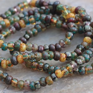 May include: A strand of small, round glass beads featuring a variegated colour palette of deep forest green, earthy brown, amber, and teal. The beads have a glossy, semi-translucent finish with subtle variations in tone and texture throughout the strand.