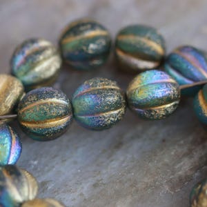 Peut inclure: Un fil de perles de verre iridescentes. Les perles sont rondes avec une texture côtelée et une bande dorée. Les couleurs varient du teal à l'or et au violet. Les perles sont enfilées sur une épingle métallique.