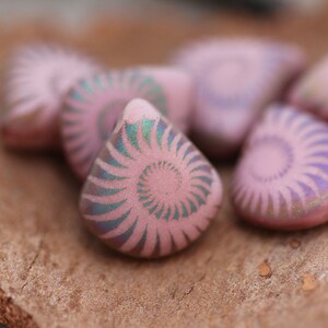 May include: A group of six pink ceramic beads with a spiral design. The beads have a iridescent finish and are arranged on a brown surface.