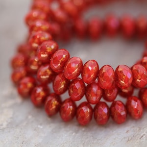 May include: Close-up of a strand of red, faceted beads with a golden shimmer. The oval-shaped beads are arranged in a circular pattern, possibly a necklace or bracelet. The background is a blurred, neutral tone.