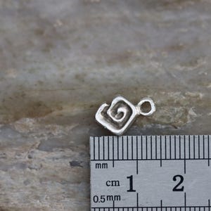 Puede incluir: Un amuleto de plata con un dise&ntilde;o en espiral, que mide aproximadamente 1 cm de di&aacute;metro. El amuleto se muestra junto a una regla para referencia de tama&ntilde;o.