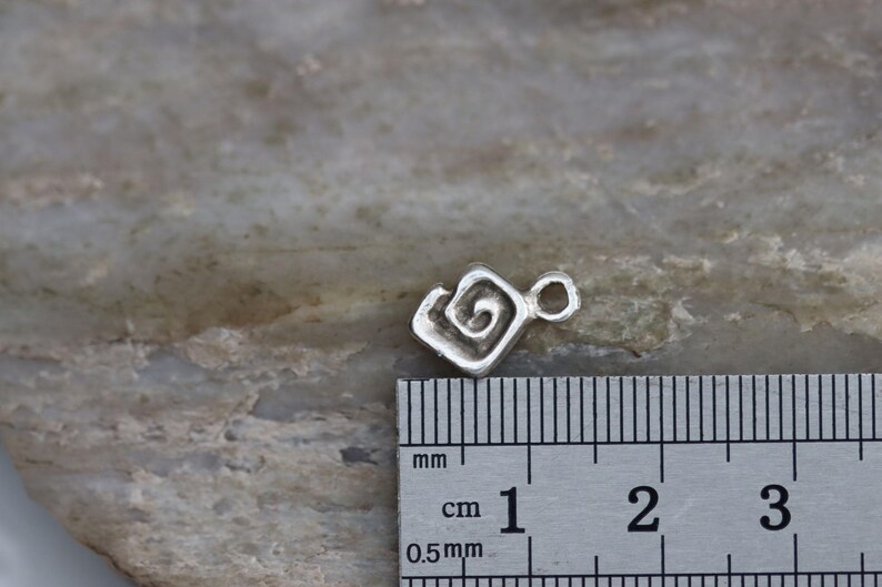 Puede incluir: Un amuleto de plata con un dise&ntilde;o en espiral, que mide aproximadamente 1 cm de di&aacute;metro. El amuleto se muestra junto a una regla para referencia de tama&ntilde;o.