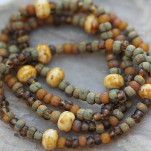 May include: A multi-strand necklace made of small, brown, green, and white glass beads. The beads are arranged in a random pattern, creating a unique and eye-catching design.