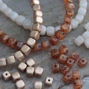 May include: A collection of square beads in three distinct finishes: matte metallic gold, translucent white, and brown marbled. The beads are arranged in loose strands and scattered individual pieces, each featuring a central hole for threading.