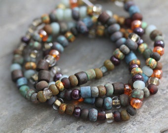 May include: A multi-strand necklace made of small, colorful beads. The beads are in shades of brown, blue, green, and orange. The necklace is made of a variety of materials, including glass, metal, and wood.