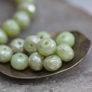 May include: A spoon filled with light green faceted glass beads. The beads have a subtle iridescent sheen.