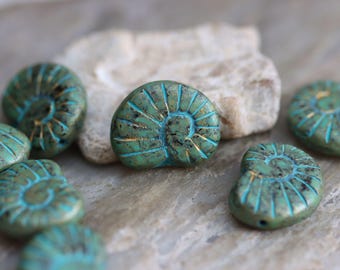 May include: A collection of turquoise-colored, shell-shaped beads with gold accents. The beads have a spiral design and are arranged on a neutral-toned surface. The beads are likely for jewelry making or other craft projects.