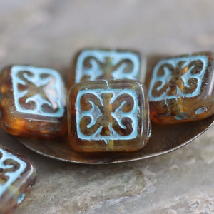 May include: Six square glass beads with a light blue design on a brown background. The beads are on a silver spoon.