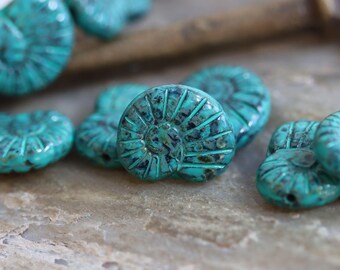 May include: Close-up of several turquoise-colored, fossil-shaped beads. Each bead features a detailed spiral design with dark blue accents. The beads are scattered on a textured, light brown surface, suggesting a craft or jewelry-making theme.