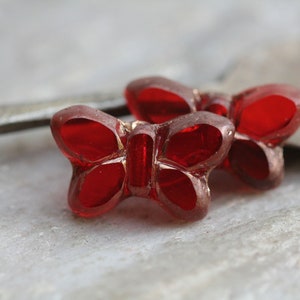 May include: Two red glass butterfly shaped beads. The beads are translucent and have a shiny finish.