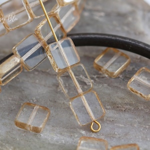May include: A strand of clear glass square beads with gold accents. The beads are strung on a gold wire and there are loose beads on a grey surface.