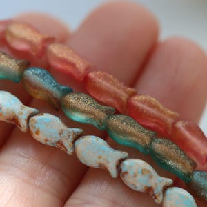 以下が含まれることがあります： さまざまな色の魚の形をしたビーズの3つのストランドのクローズアップ。一番上のストランドは半透明のコーラル色、真ん中のストランドはティールとゴールド、一番下のストランドはライトブルーとブラウンのアクセントです。ビーズは一緒にひもでつながれています。