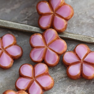 Puede incluir: Una colección de cuentas con forma de flor que presentan un centro rosa brillante con cinco pétalos redondeados y un borde liso de color bronce metálico. Cada cuenta tiene un diseño distintivo de cinco pétalos con un centro elevado.