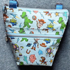 May include: A light blue pouch with a zip closure, decorated with a cartoon pattern of characters such as a green dinosaur and a cowboy. The pouch has two zipped sections and blue straps.