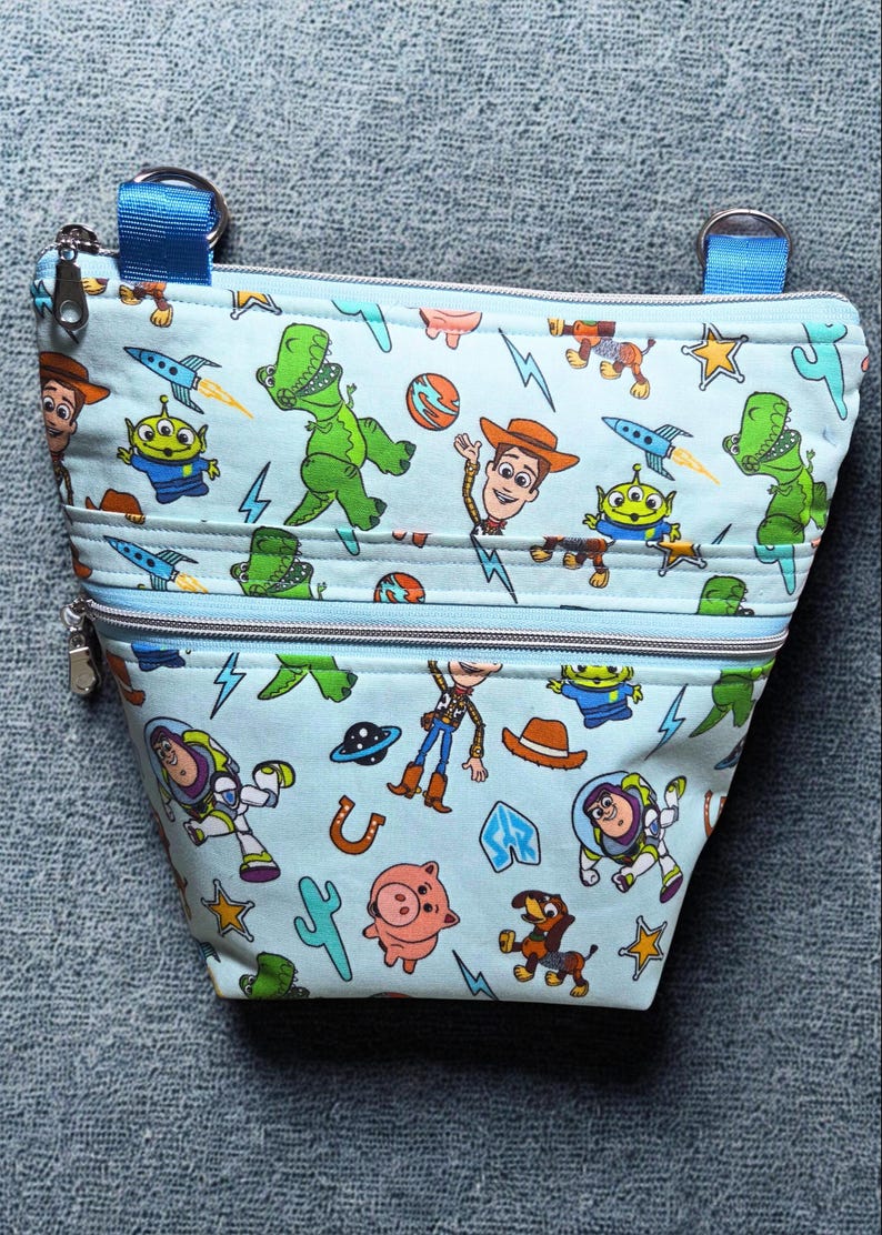 May include: A light blue pouch with a zip closure, decorated with a cartoon pattern of characters such as a green dinosaur and a cowboy. The pouch has two zipped sections and blue straps.
