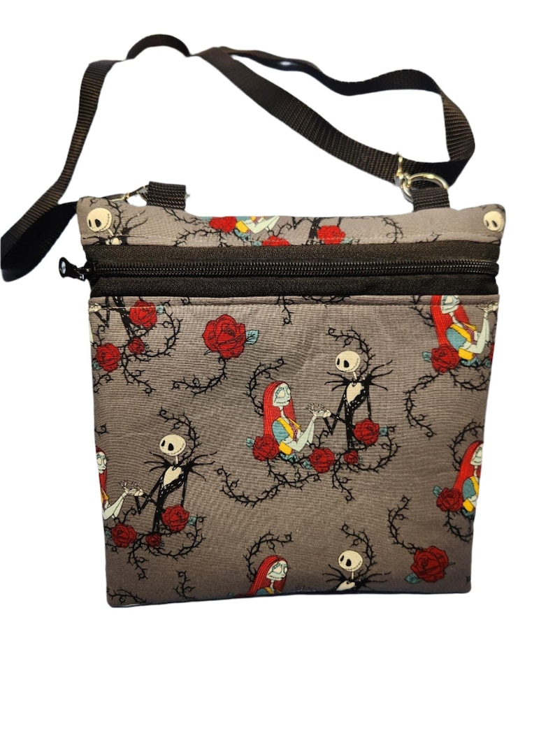 May include: A gray crossbody bag with a black strap and zipper closure. The bag features a repeating pattern of Jack Skellington and Sally from The Nightmare Before Christmas, with red roses and thorny vines.
