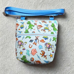 May include: A light blue crossbody bag with a colourful print of Toy Story characters. It has a blue zip, an adjustable blue strap, and silver hardware. The bag is designed for carrying small items, with a strap length that can be adjusted.