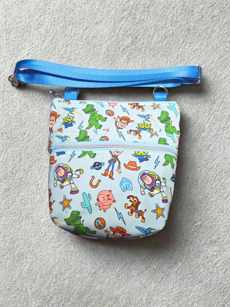 May include: A light blue crossbody bag with a colourful print of Toy Story characters. It has a blue zip, an adjustable blue strap, and silver hardware. The bag is designed for carrying small items, with a strap length that can be adjusted.