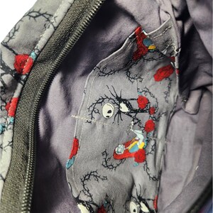 May include: A gray zippered bag with a black zipper. The inside of the bag is lined with a gray fabric featuring a repeating pattern of black vines, red roses, and a character from the Nightmare Before Christmas.