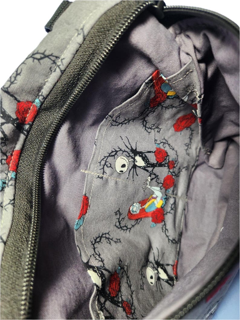 May include: A gray zippered bag with a black zipper. The inside of the bag is lined with a gray fabric featuring a repeating pattern of black vines, red roses, and a character from the Nightmare Before Christmas.