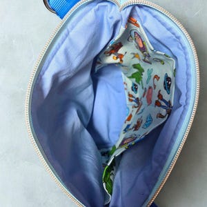 May include: An open, light blue zipped bag with a blue strap and gold-coloured hardware. The interior is light blue with a patterned pocket featuring cartoon characters. The zip is gold-coloured.