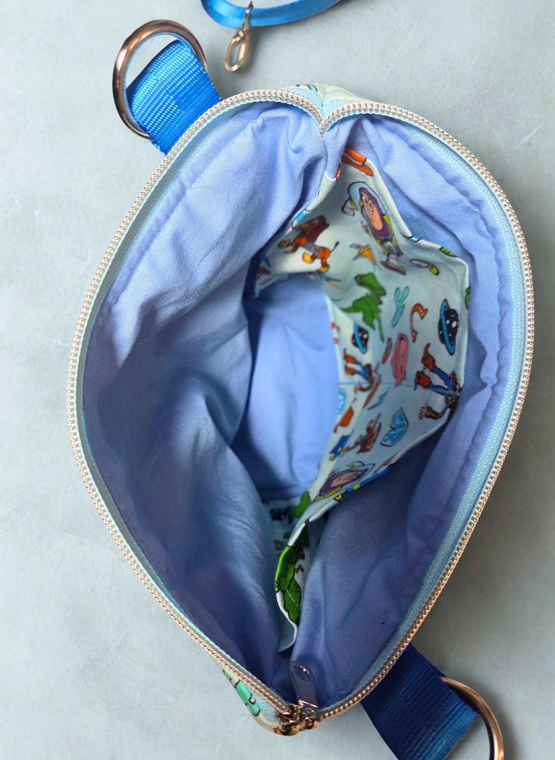 May include: An open, light blue zipped bag with a blue strap and gold-coloured hardware. The interior is light blue with a patterned pocket featuring cartoon characters. The zip is gold-coloured.