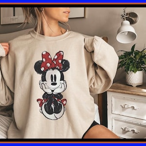 May include: A beige crewneck sweatshirt featuring a distressed graphic print of Minnie Mouse on the front. The character is depicted in black and white with a red and white polka dot bow, resting her chin on her hands in a seated position.