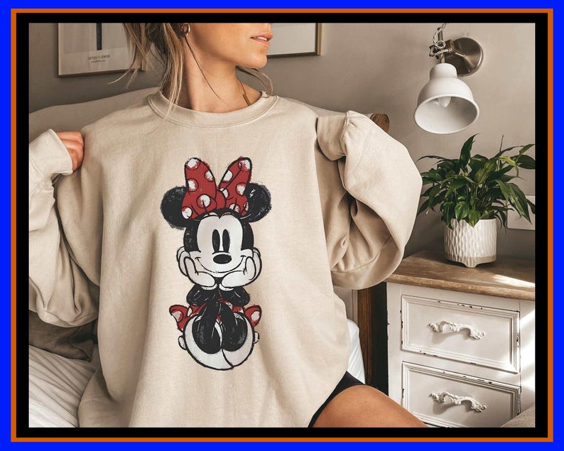 May include: A beige crewneck sweatshirt featuring a distressed graphic print of Minnie Mouse on the front. The character is depicted in black and white with a red and white polka dot bow, resting her chin on her hands in a seated position.