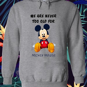 May include: A gray hooded sweatshirt featuring a graphic of Mickey Mouse. The text "WE ARE NEVER TOO OLD FOR MICKEY MOUSE" is printed above and below the character. The design includes Mickey's iconic ears, red shorts, and yellow shoes.