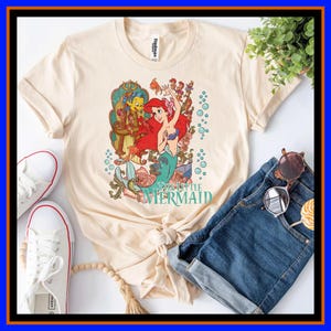 May include: A cream-colored t-shirt featuring a graphic of Ariel from The Little Mermaid with Flounder, surrounded by floral elements and bubbles. The text The Little Mermaid is printed in teal below the character illustration on the front.