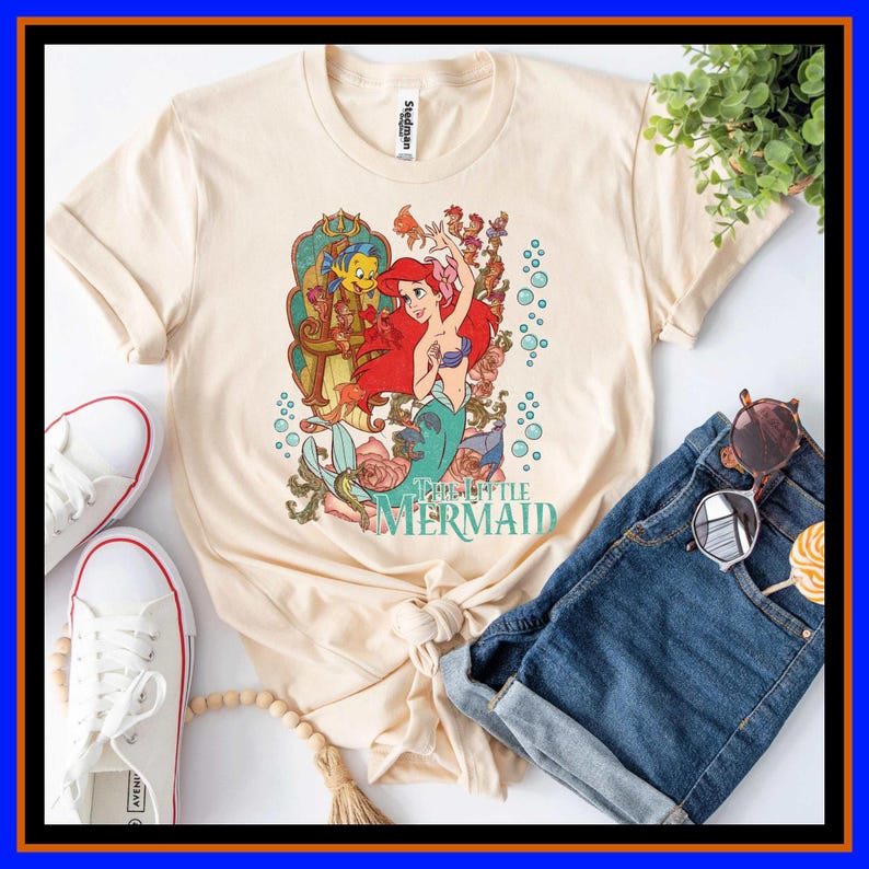 May include: A cream-colored t-shirt featuring a graphic of Ariel from The Little Mermaid with Flounder, surrounded by floral elements and bubbles. The text The Little Mermaid is printed in teal below the character illustration on the front.
