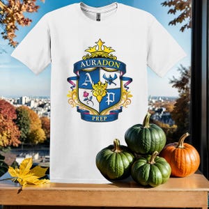 May include: White t-shirt with a blue and gold crest featuring the words "Auradon Prep" and a crown. The crest has a blue shield with a letter "A" and a letter "P" on it. There are pumpkins in the background.