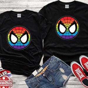Puede incluir: Dos camisetas negras con un logotipo de Spiderman de colores arco&iacute;ris. El logotipo es un c&iacute;rculo con una telara&ntilde;a y los ojos de Spiderman. Las camisetas est&aacute;n sobre una superficie de madera con un par de zapatillas rojas.