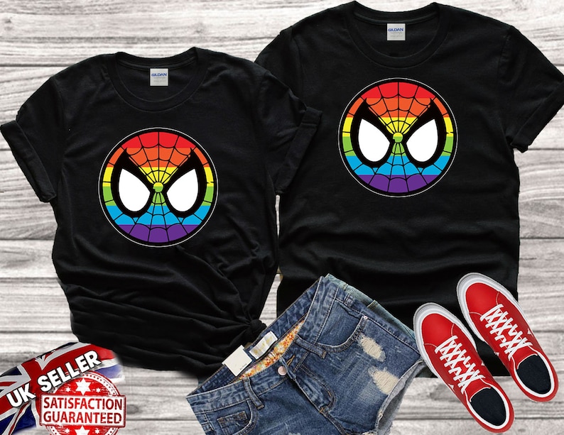 Puede incluir: Dos camisetas negras con un logotipo de Spiderman de colores arco&iacute;ris. El logotipo es un c&iacute;rculo con una telara&ntilde;a y los ojos de Spiderman. Las camisetas est&aacute;n sobre una superficie de madera con un par de zapatillas rojas.
