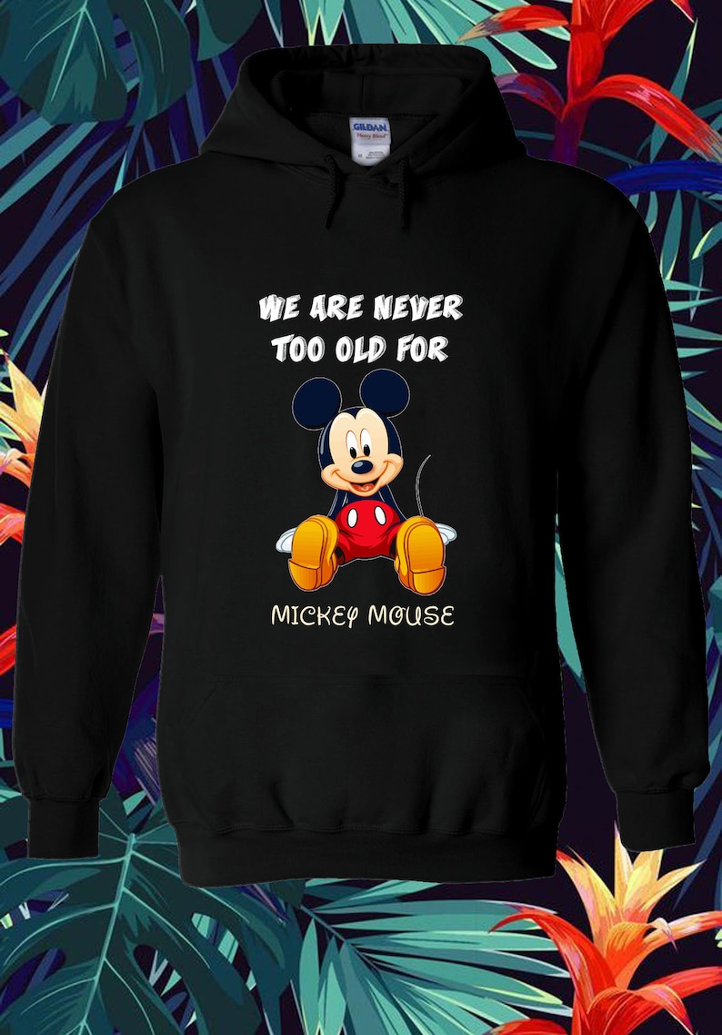 May include: Black hooded sweatshirt featuring a seated Mickey Mouse graphic. The text "WE ARE NEVER TOO OLD FOR MICKEY MOUSE" is printed above and below the character. The design includes Mickey's signature red shorts, yellow shoes, and black ears.