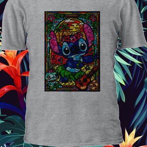 Pu&ograve; includere: Una maglietta grigia con un'immagine di Stitch di Lilo & Stitch in stile vetrata. Stitch sta suonando un ukulele e indossa una collana di fiori. L'immagine &egrave; circondata da una cornice colorata.