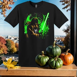 Puede incluir: Una camiseta negra con un gráfico verde y blanco de un personaje de Star Wars blandiendo una espada láser. El personaje está rodeado de un fondo verde y blanco similar a una galaxia.